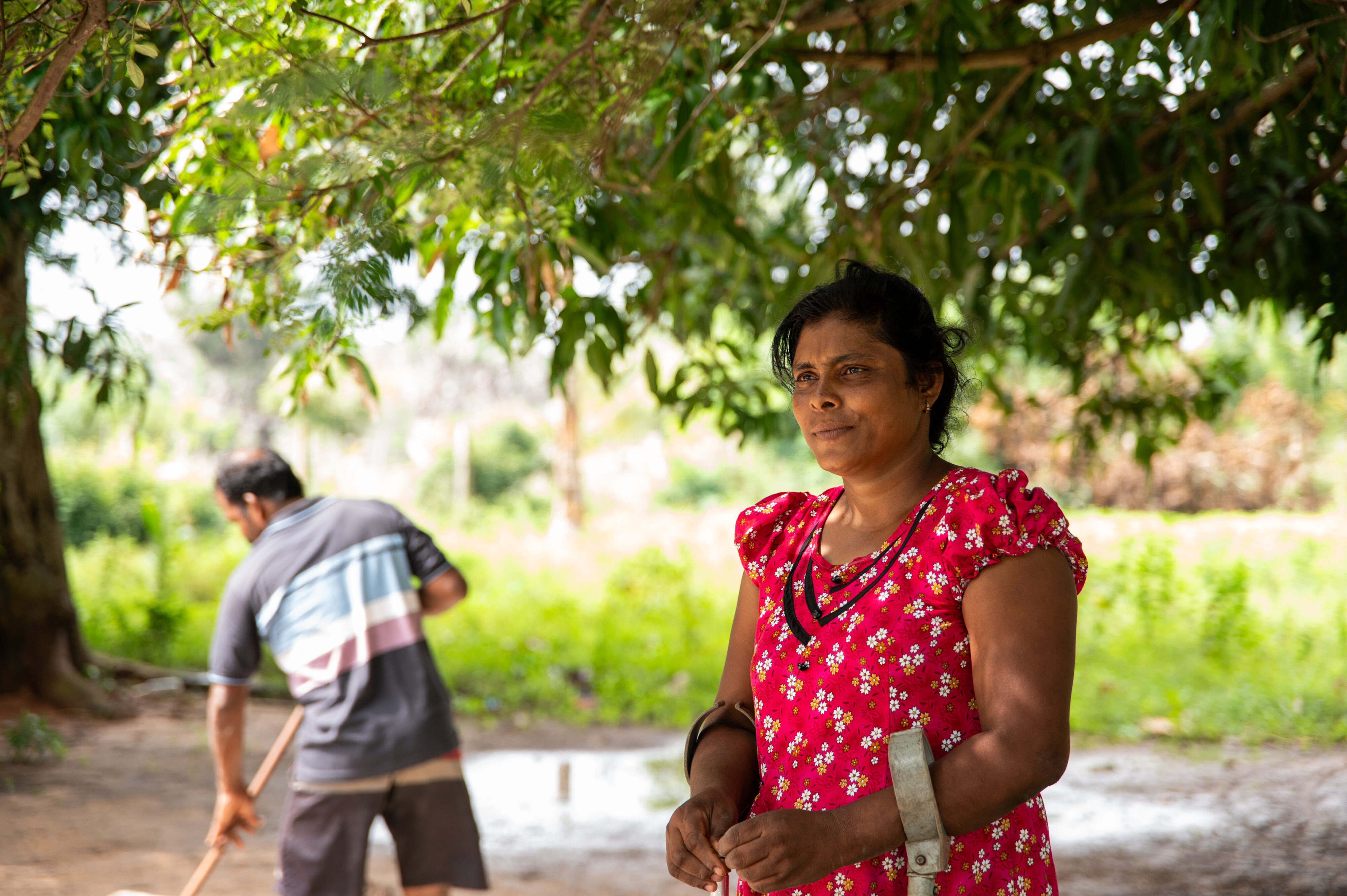 Woman farmer standing under a tree while another person works in the field behind her.
