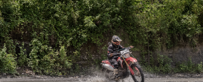 Motorcyclist riding a dirt bike through a shallow stream surrounded by green forest.
