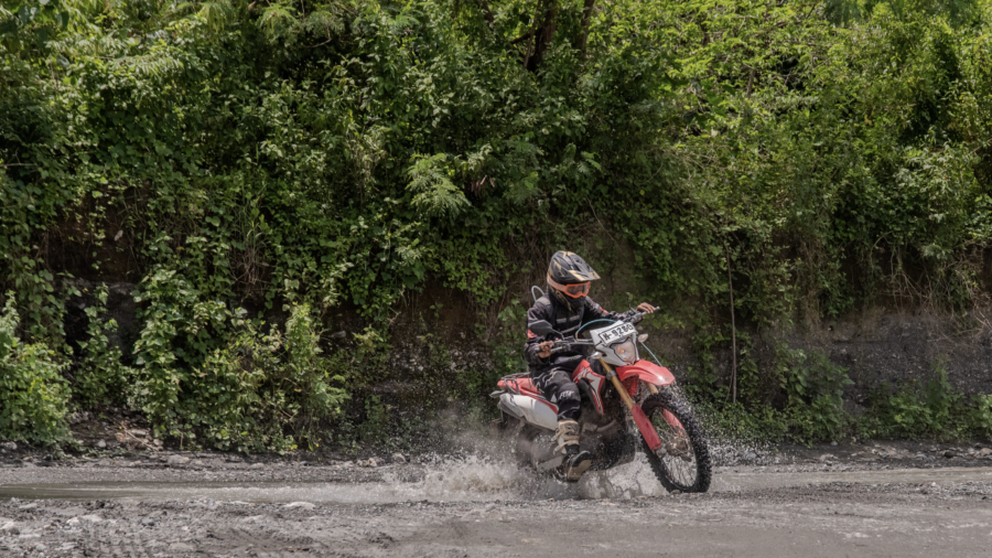 Motorcyclist riding a dirt bike through a shallow stream surrounded by green forest.