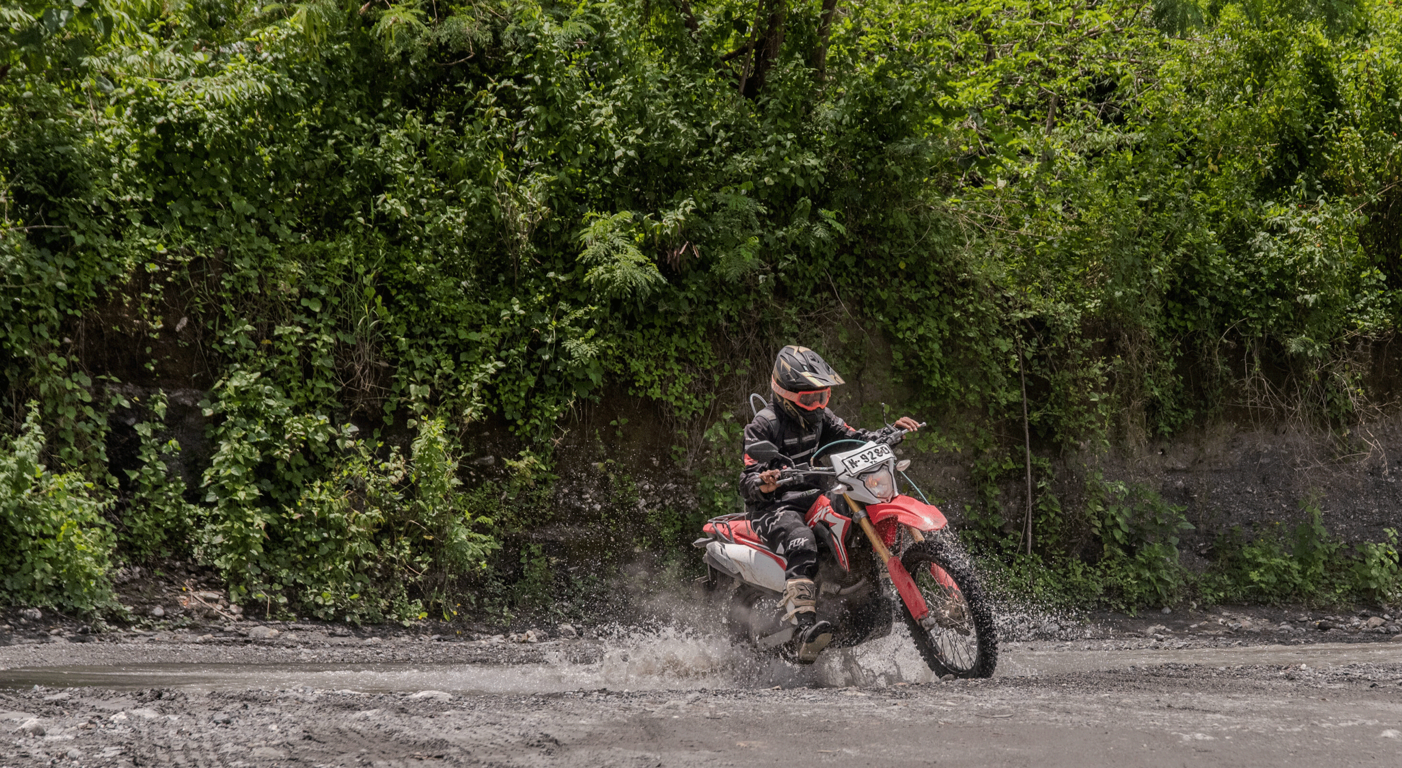 Motorcyclist riding a dirt bike through a shallow stream surrounded by green forest.