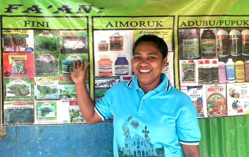 Woman standing in front of agricultural product posters at a farm input shop in Timor-Leste.