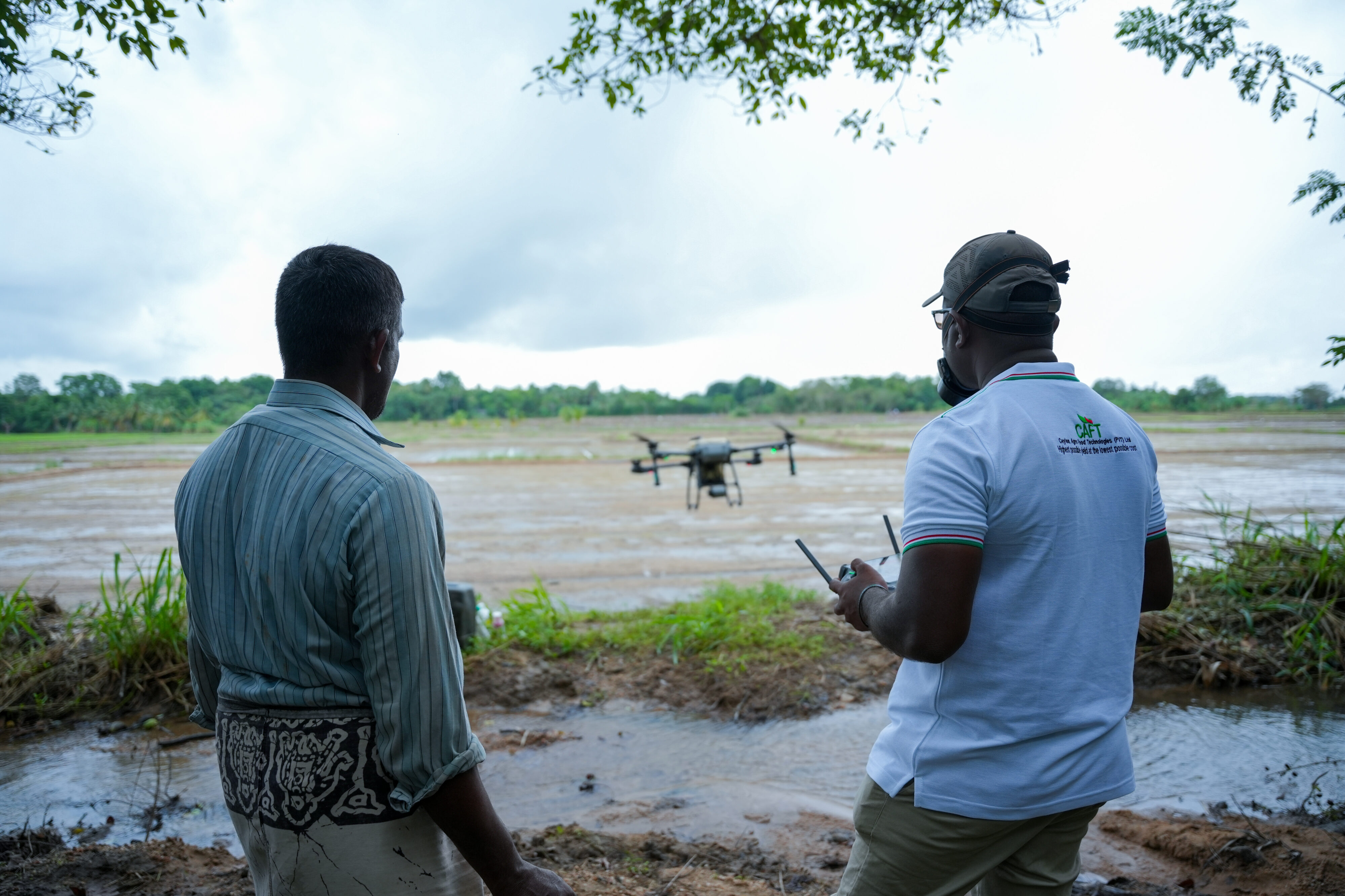 Two men watching a drone fly over a flooded agricultural field in Sri Lanka.
