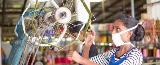 A woman wearing a face mask spins yarn on a wheel, highlighting the traditional craftsmanship of Selyn Handlooms in Sri Lanka.