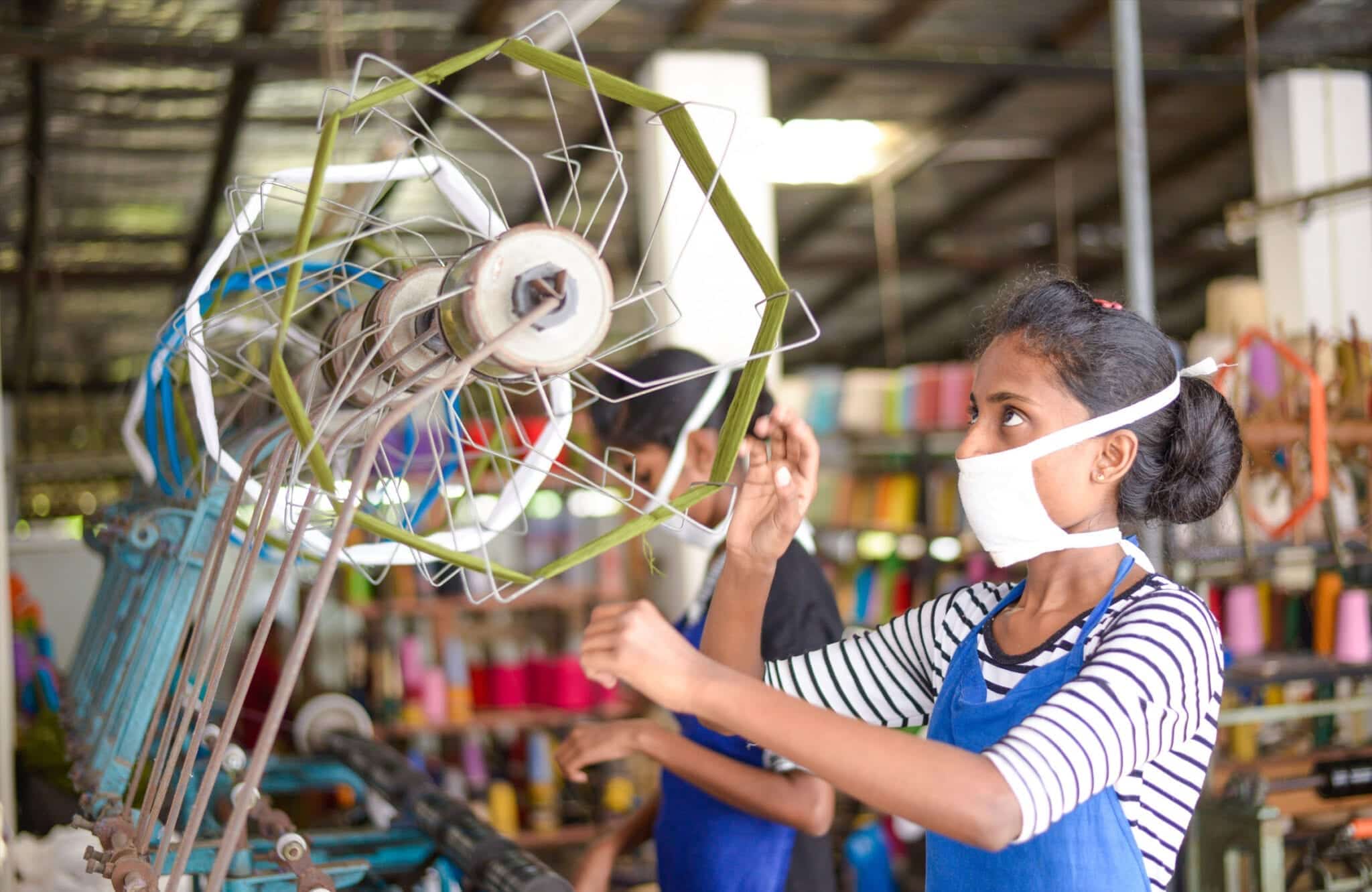 A woman wearing a face mask spins yarn on a wheel, highlighting the traditional craftsmanship of Selyn Handlooms in Sri Lanka.